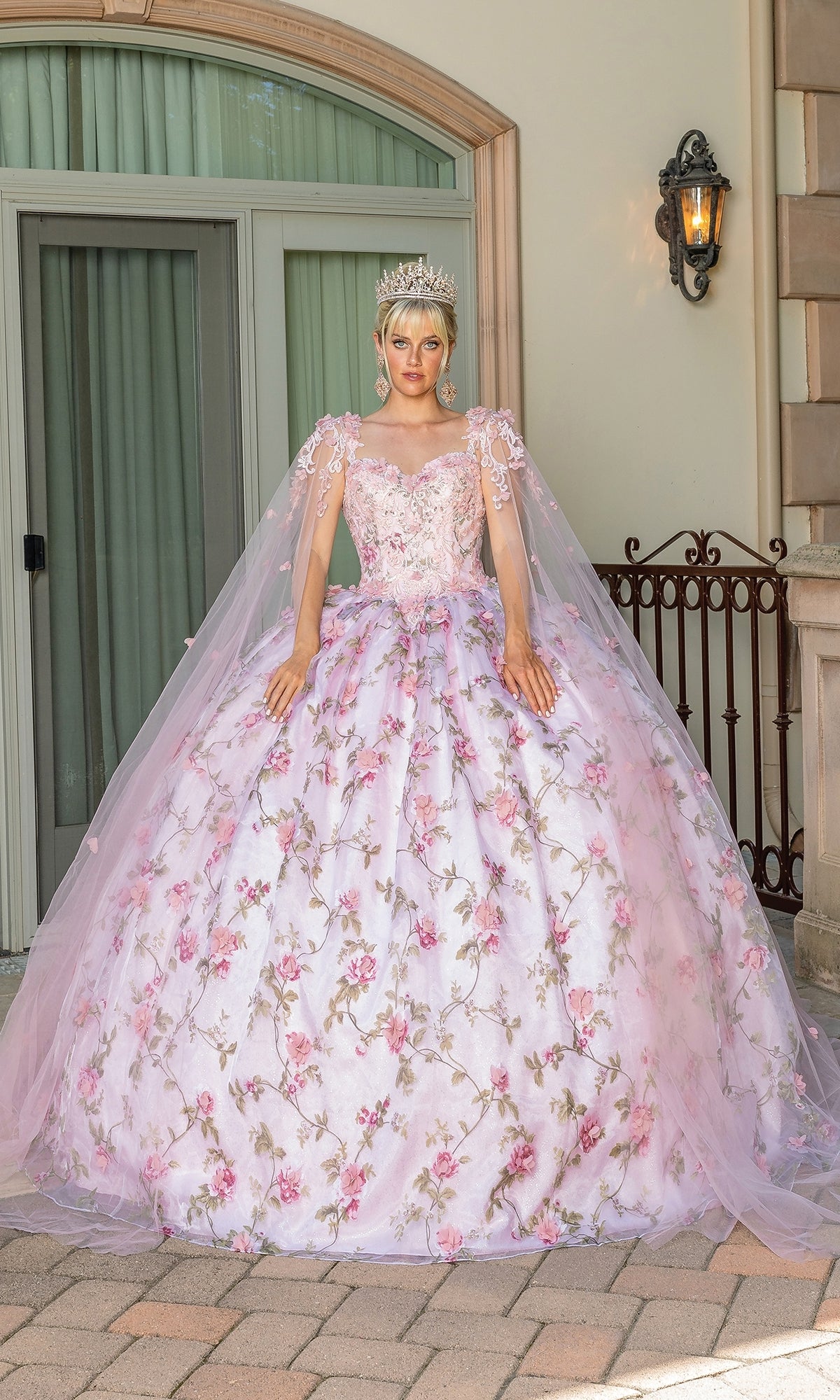 A person stands outdoors in front of large windows and a lantern, wearing the Quinceanera Dress 1715 By Dancing Queen—a blush pink ball gown with lace detailing—paired with a tiara.